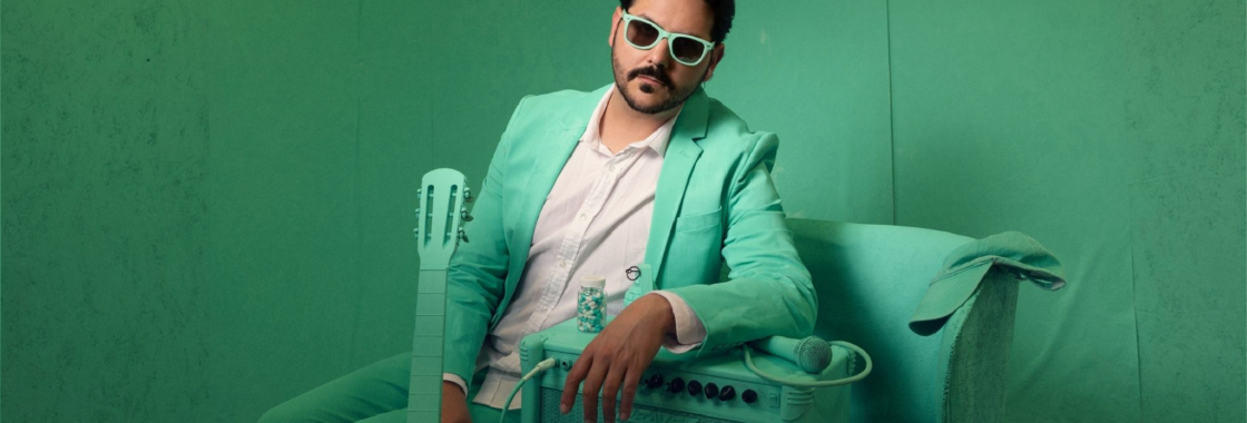 Comedian Ignacio Lopez sitting on an armchair, resting his arm on an amp, sitting in a turquoise room.